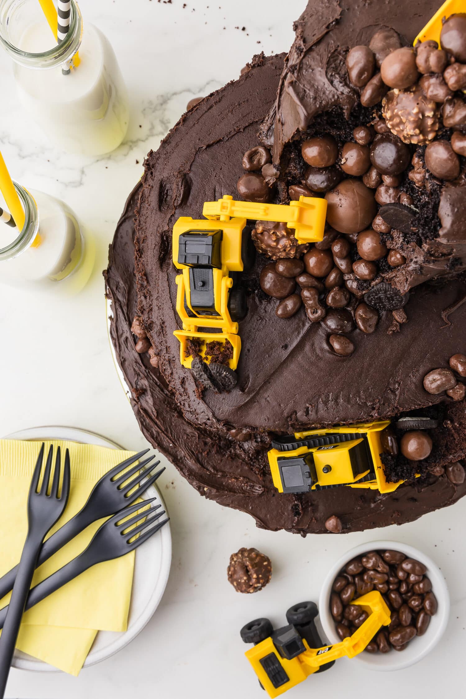 overhead photo of a double tier chocolate cake with CAT construction toys and chocolate candy boulders on a white counter with milk in bottles and yellow and black and white straws