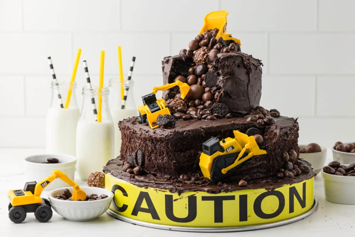 double tier chocolate cake with CAT construction toys and chocolate candy boulders on a white counter with milk in bottles and yellow and black and white straws