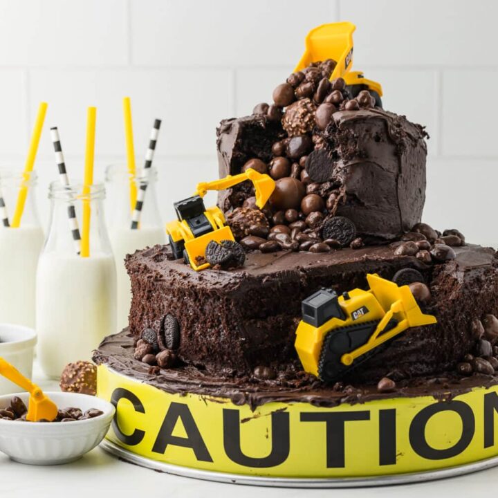 Construction cake made from two tiers of chocolate ckae with a ramp for a bulldozer, a dump truck, and a backhoe, on a white counter with cute milk jugs and construction themed drinking straws
