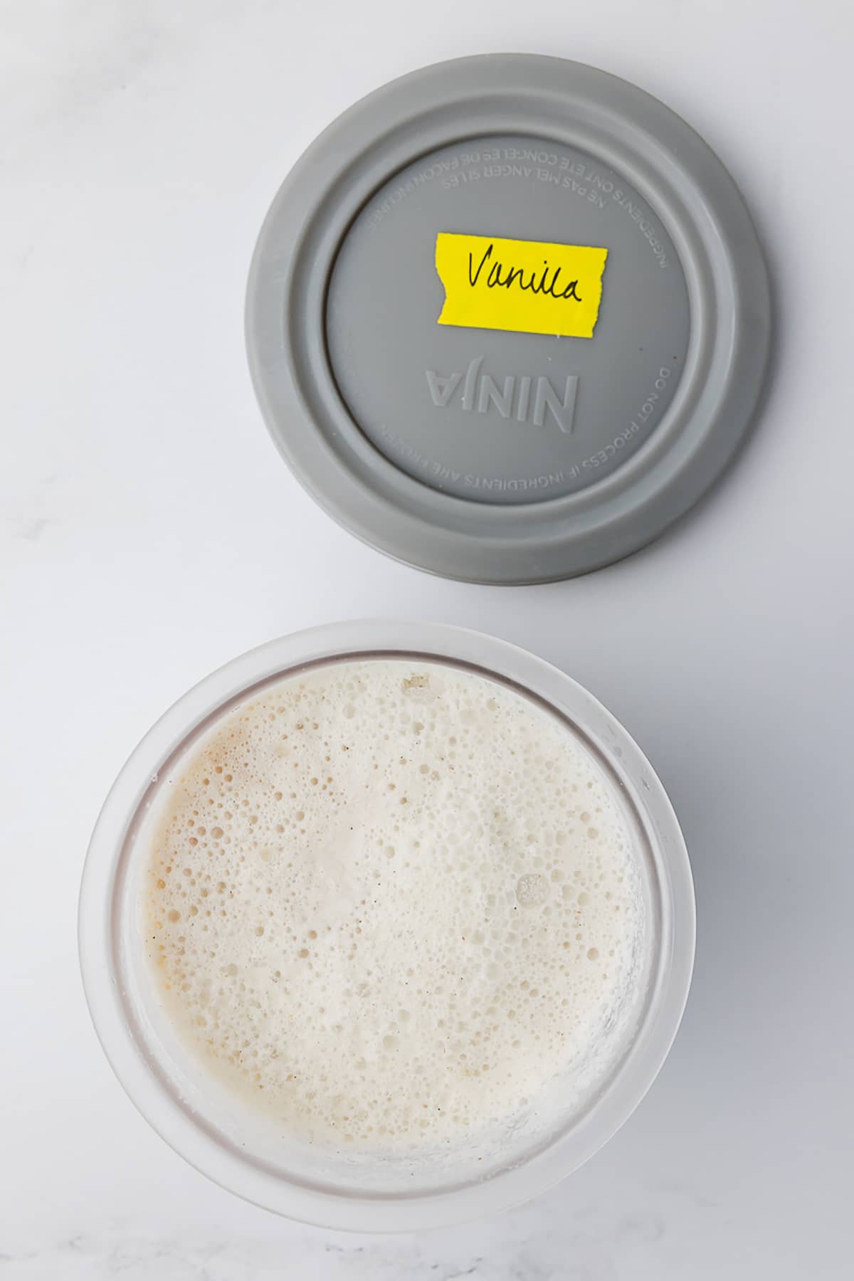 frozen vanilla ice cream in a ninja creami pot, the lid has vanilla written on a yellow piece of tape