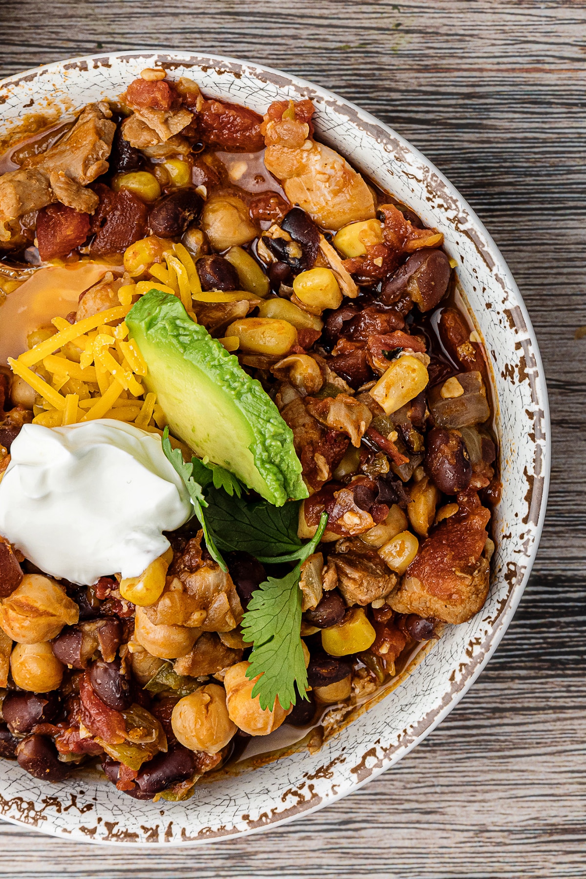 bowl of crock pot chicken chili on a wooden table with shredded cheddar cheese and a slice of avocado and dollop of sour cream