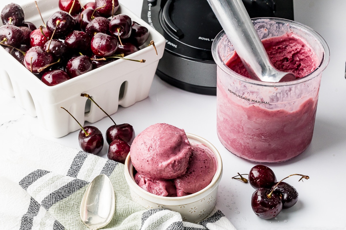 Ninja Creami Cherry Ice cream in a pint bowl with a berry bowl full of dark cherries and a bowl of ice cream on a white counter