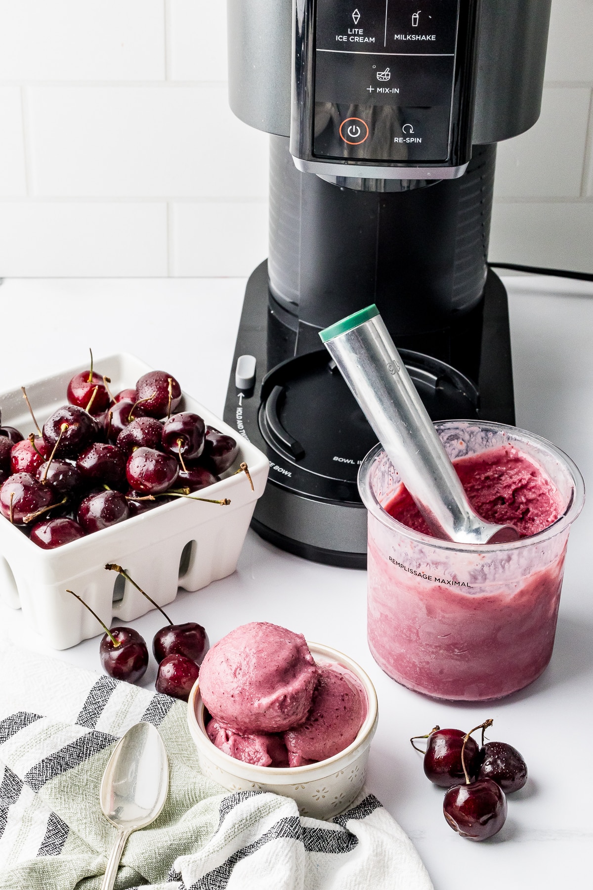 Ninja Creami Cherry Ice cream in a pint bowl with a berry bowl full of dark cherries and a bowl of ice cream on a white counter with the machine