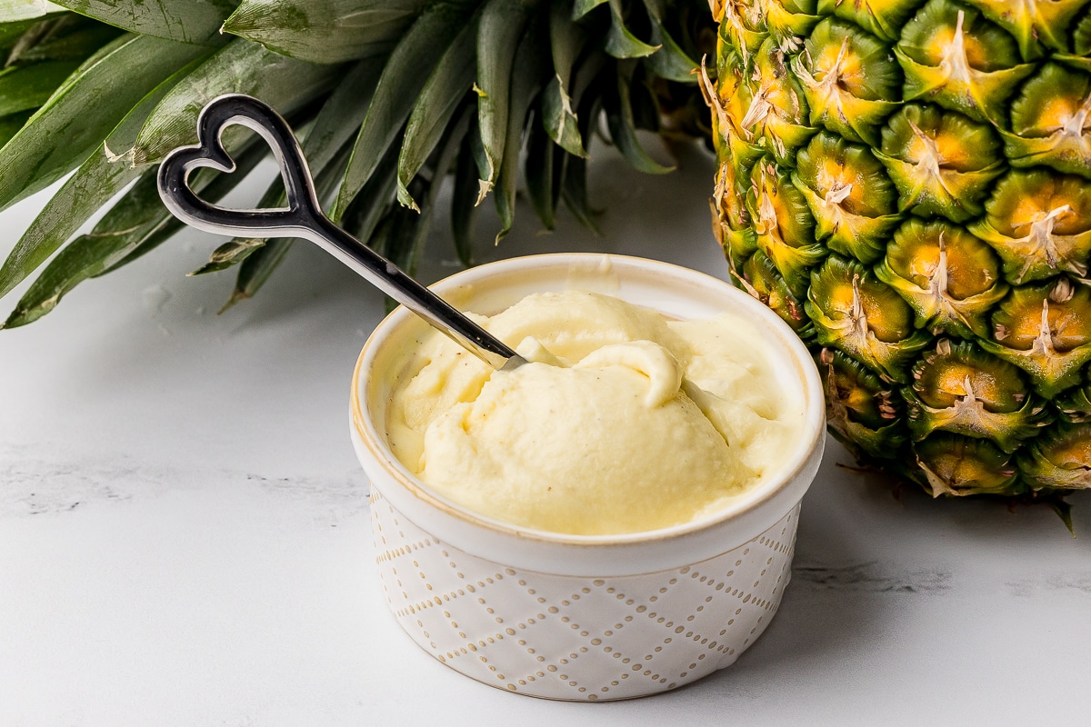 A small bowl of yellow ice cream with a silver spoon and pineapples on a counter
