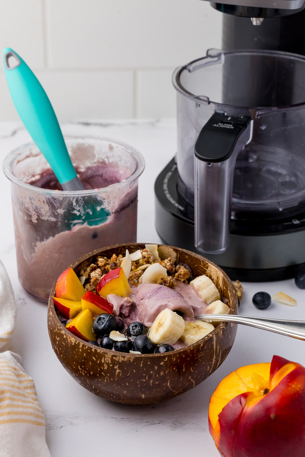 acai bowl with fresh fruit on a white countertop, nectarine, blueberries, bananas, and a ninja creami with a yelow and white kitchen towel