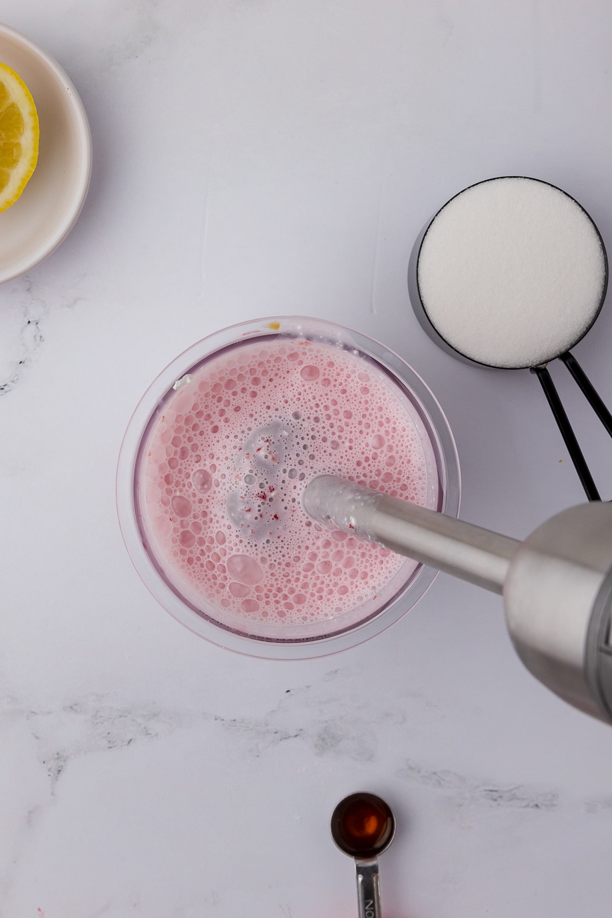 ninja creami pint jar with strawberry cheesecake mixture with an immersion blender, sugar, and vanilla on a white counter