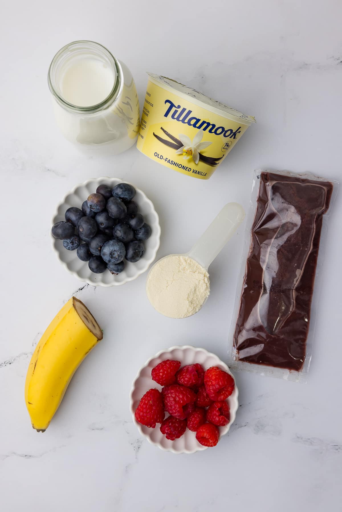 ingredients for ninja creami acai bowl including vanilla tillamook vanilla bean yogurt, milk, blueberries, protein powder, half a banana, raspberries and an acai packet