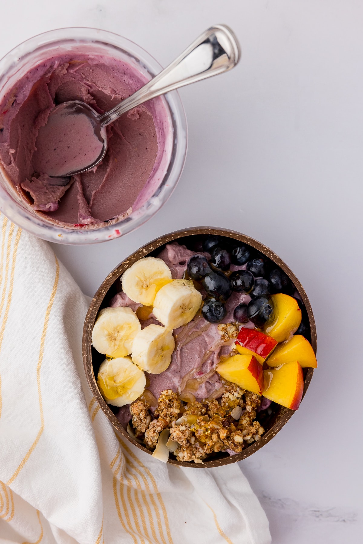 White countertop with an acai bowl with nectarines, blueberries, banana slices, and granola and a spoon, and a ninja foodie pint and a yellow striped kitchen towel.