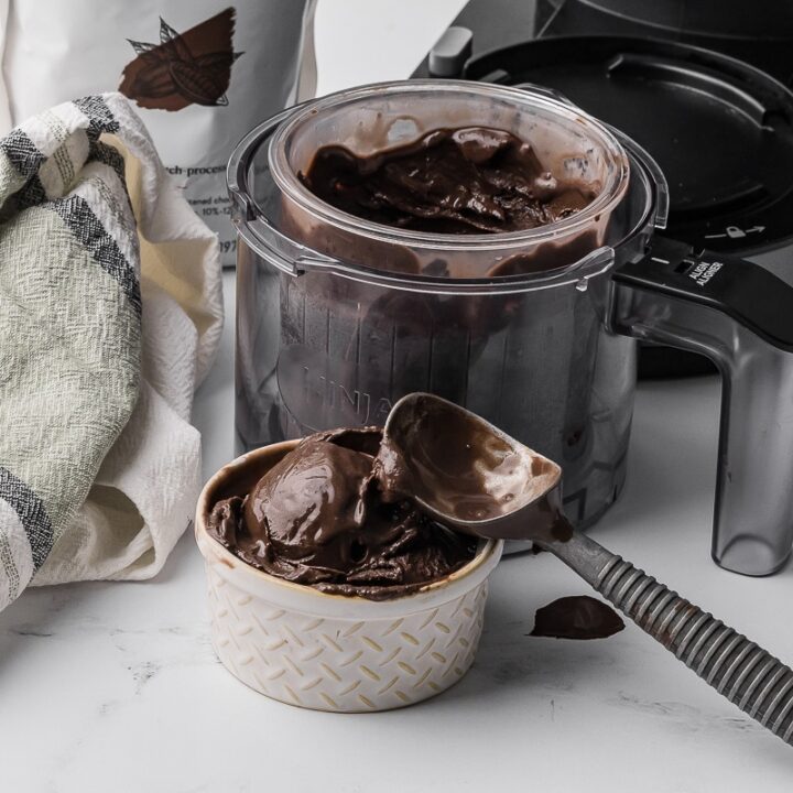 Chocolate ice cream in a small bowl with an ice cream scoop, a ninja creami bowl and a bag of dark chocolate