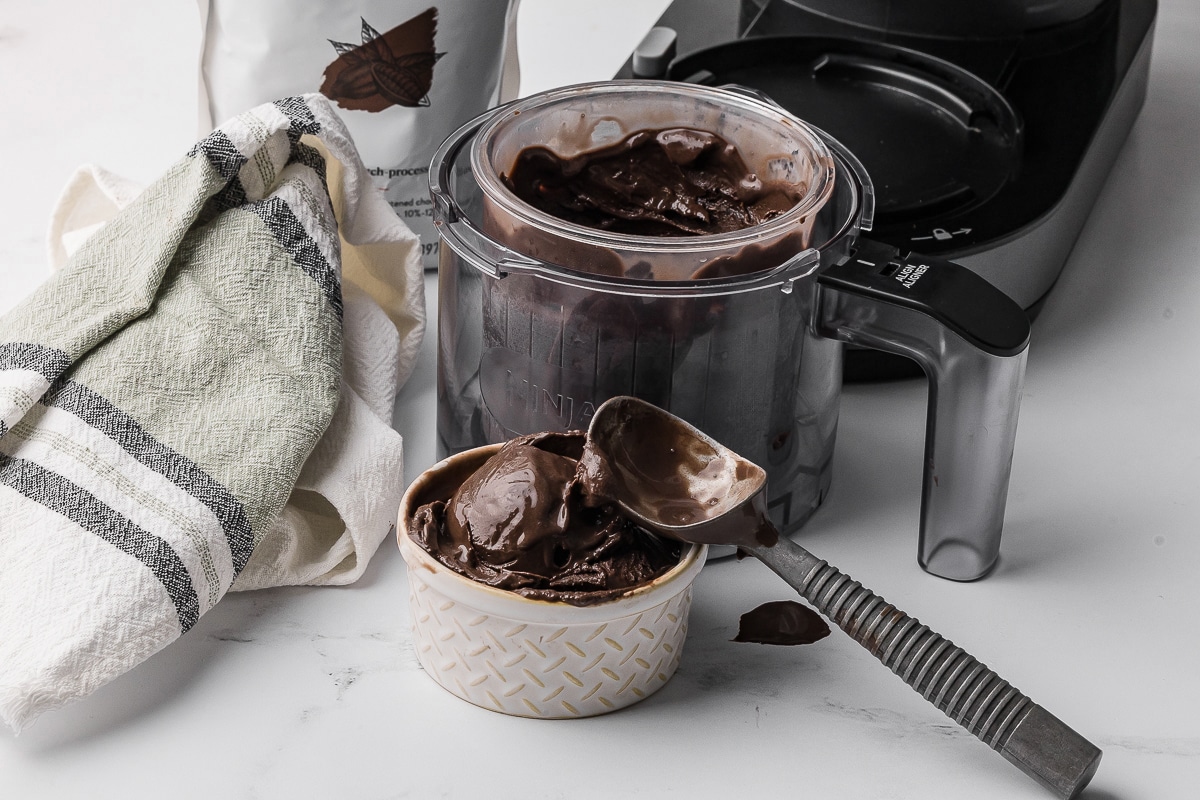 Chocolate ice cream in a small bowl with an ice cream scoop, a ninja creami bowl and a bag of dark chocolate