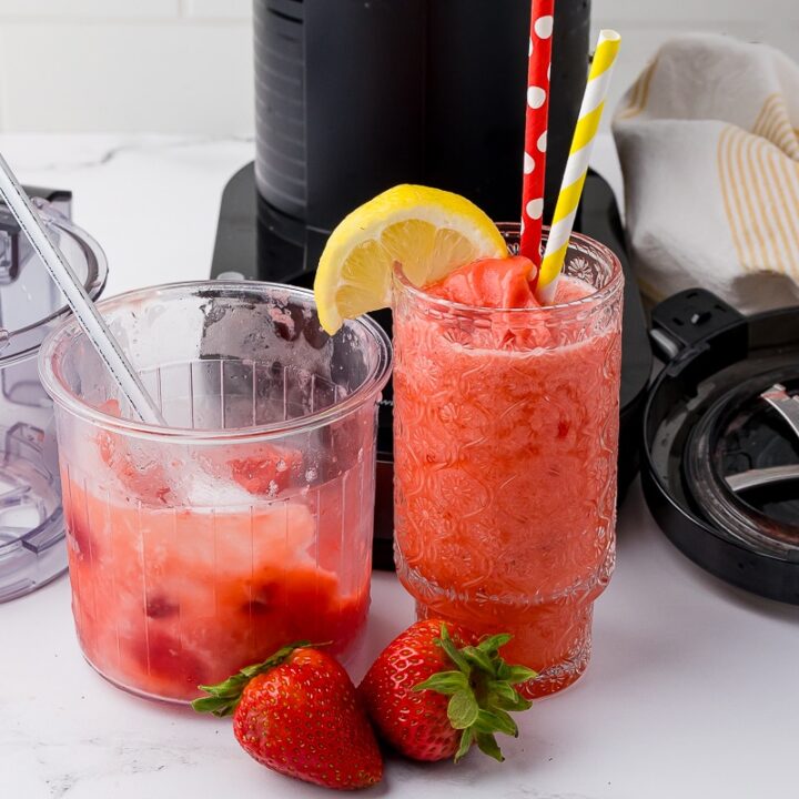 ninja creami strawberry lemonade slushie in a patterned glass with a red polka dot straw and a yellow striped straw. There are strawberries on the white counter as well as the ninja creami pint jar with more slushie in it behind the glass.