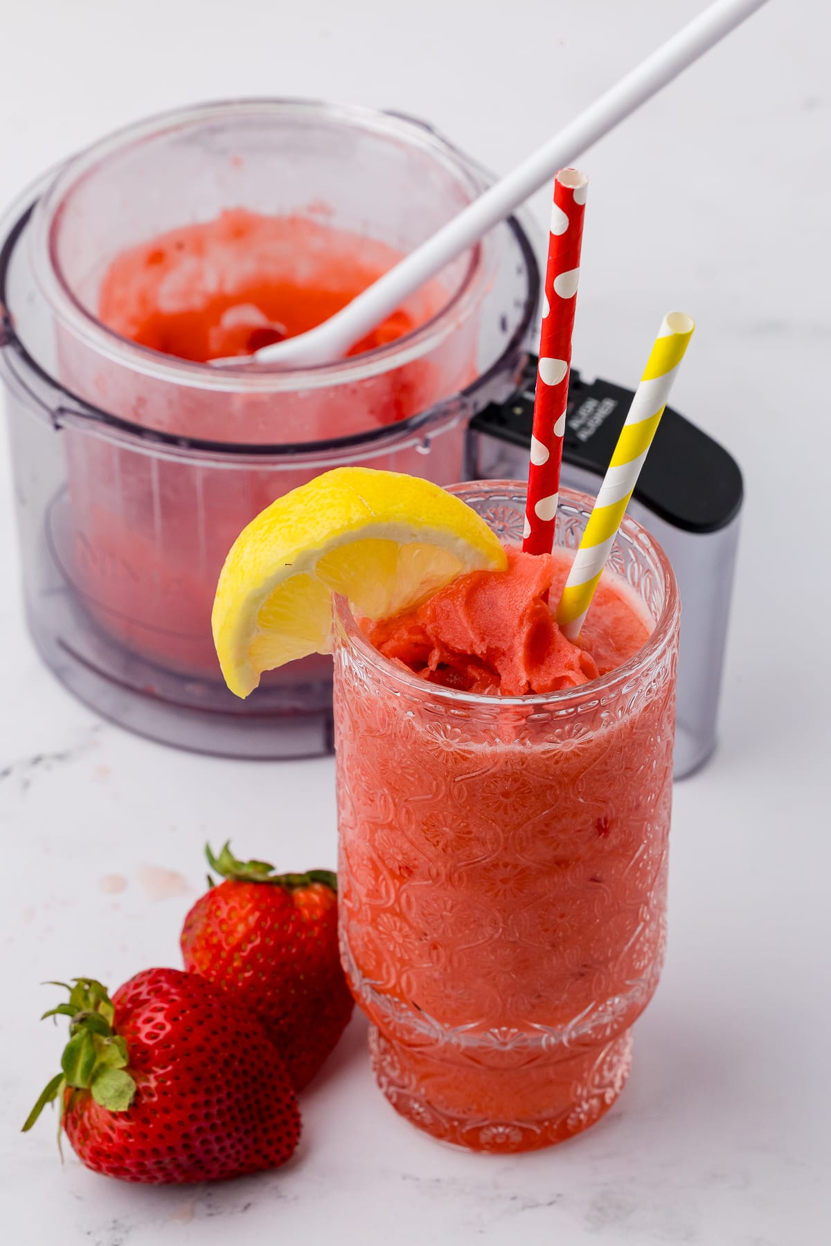 ninja creami strawberry lemonade slushie in a patterned glass with a red polka dot straw and a yellow striped straw. There are strawberries on the white counter as well as the ninja creami pint jar with more slushie in it behind the glass.