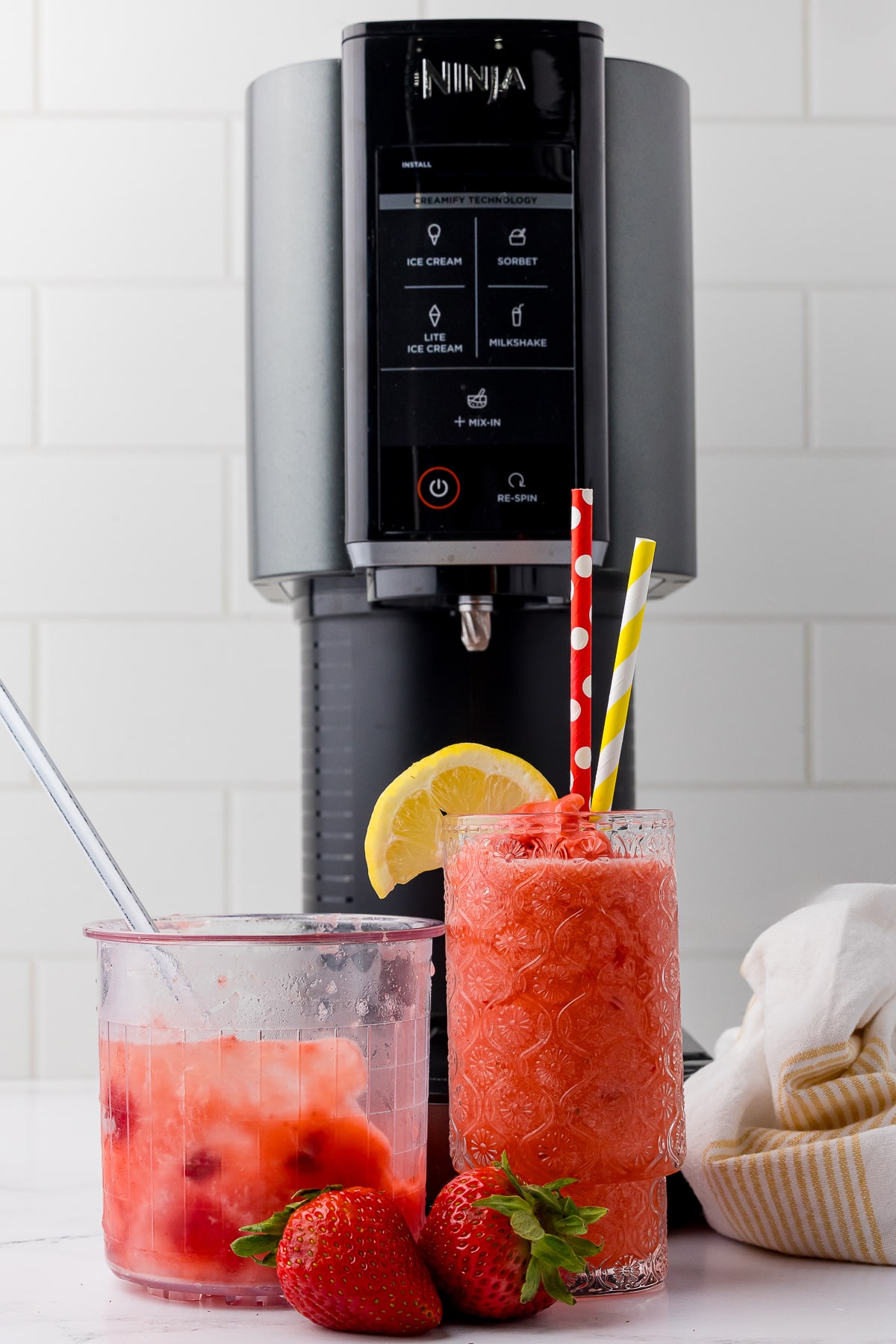 ninja creami strawberry lemonade slushie in a patterned glass with a red polka dot straw and a yellow striped straw. There are strawberries on the white counter as well as the ninja creami NC 300 behind the glass