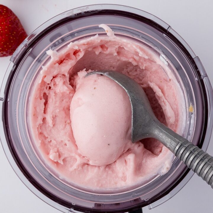 strawberry cheesecake ice cream in a ninja creami pint container with an ice cream scoop and a fresh strawberry on a white countertop