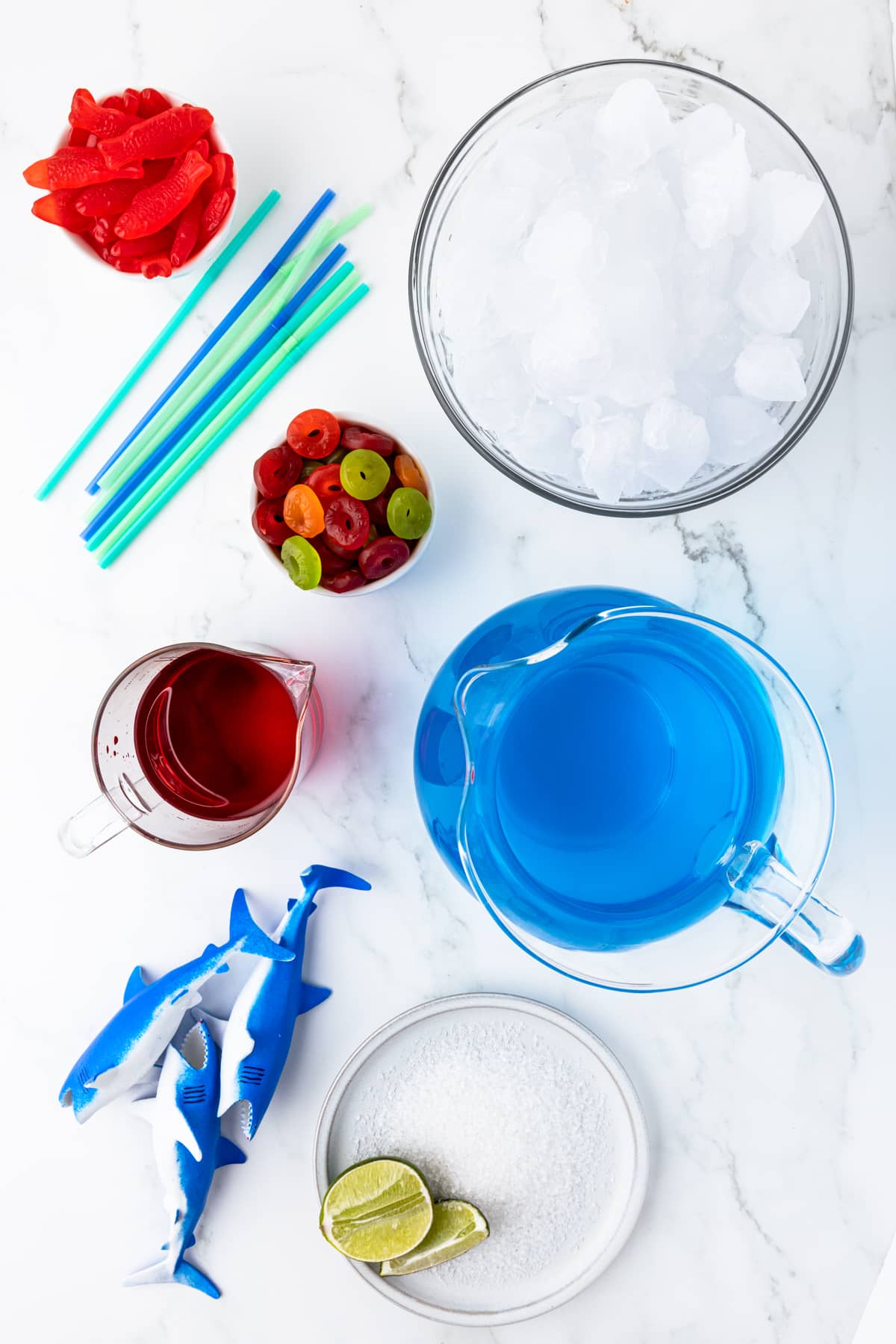 swedish fist, blue and green straws, a bowl of ice, a bowl of gummy life savers, a measuring cup of grenadine, a pitcher of blue gatorade, 3 plastic sharks, and a plate of sanding sugar with a couple of wedges of lime