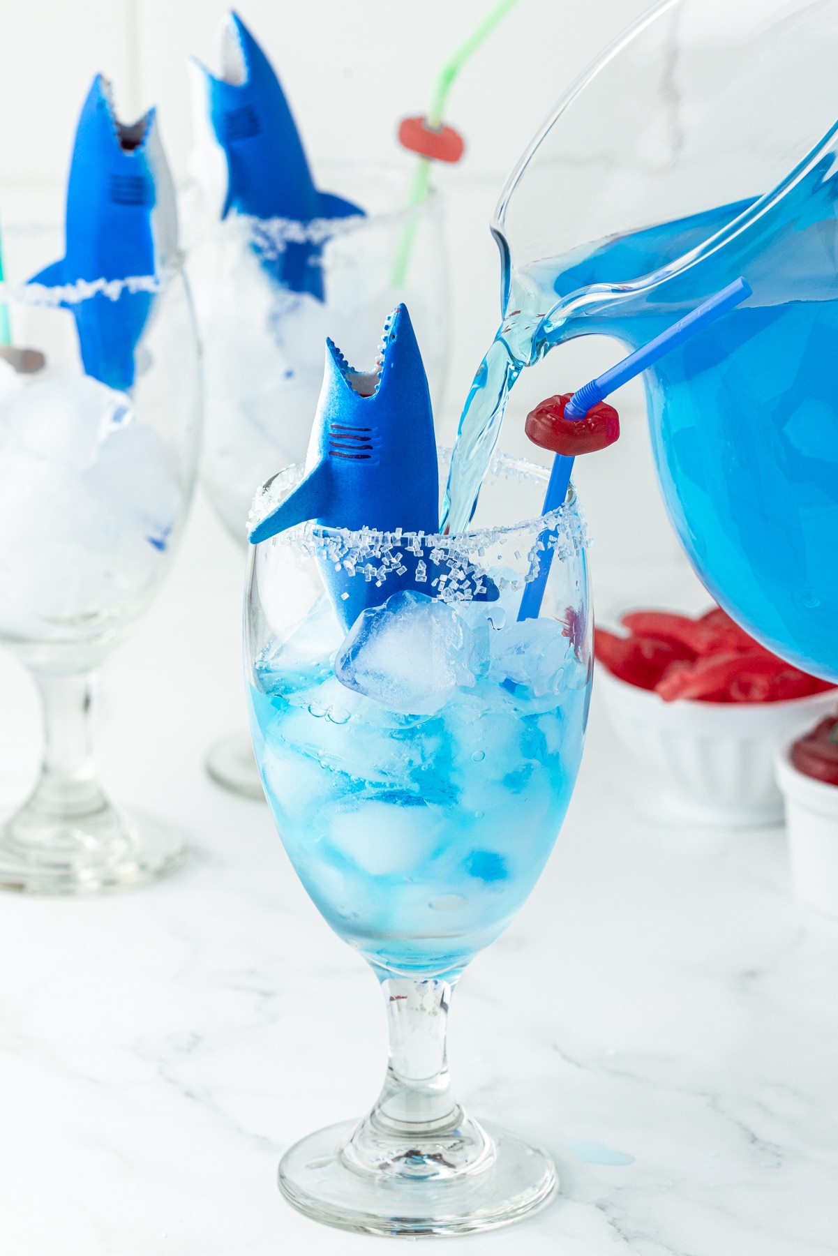 plastic toy shark in a glass filled with ice, blue gatorade being poured into the glass that has white sanding sugar on the rim