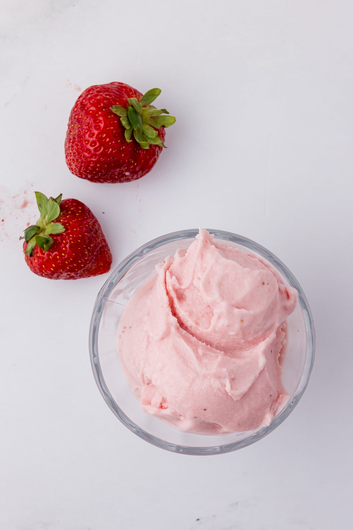 Strawberry cheesecake ice cream in a small glass bowl with two strawberries on a white counter