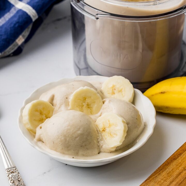 White bowl of ice cream with banana slices on the counter with a sliced banana and a blue and white towel