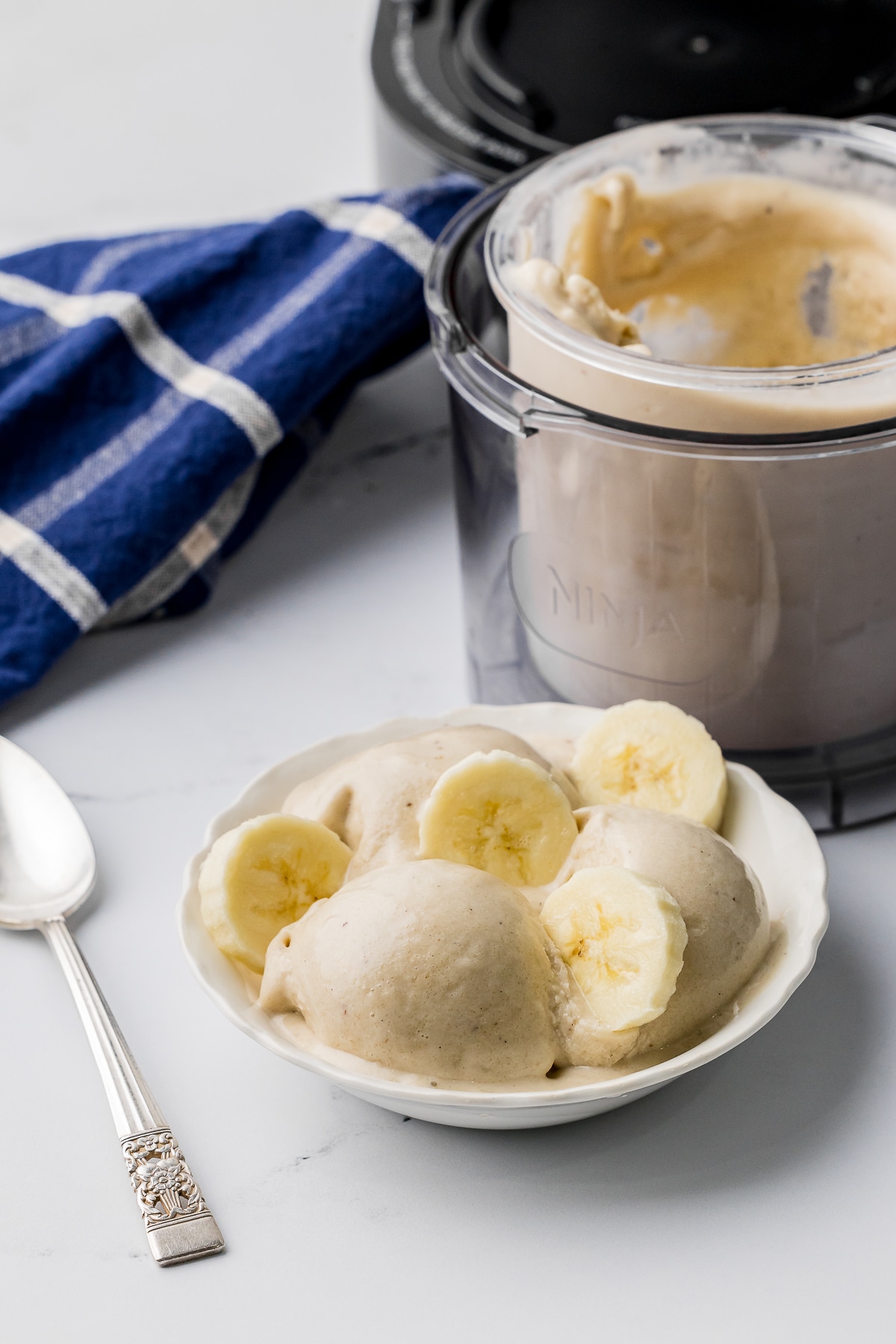 White bowl of tan colored ice cream with sliced bananas on a white counter with a Ninja Creami machine in the background