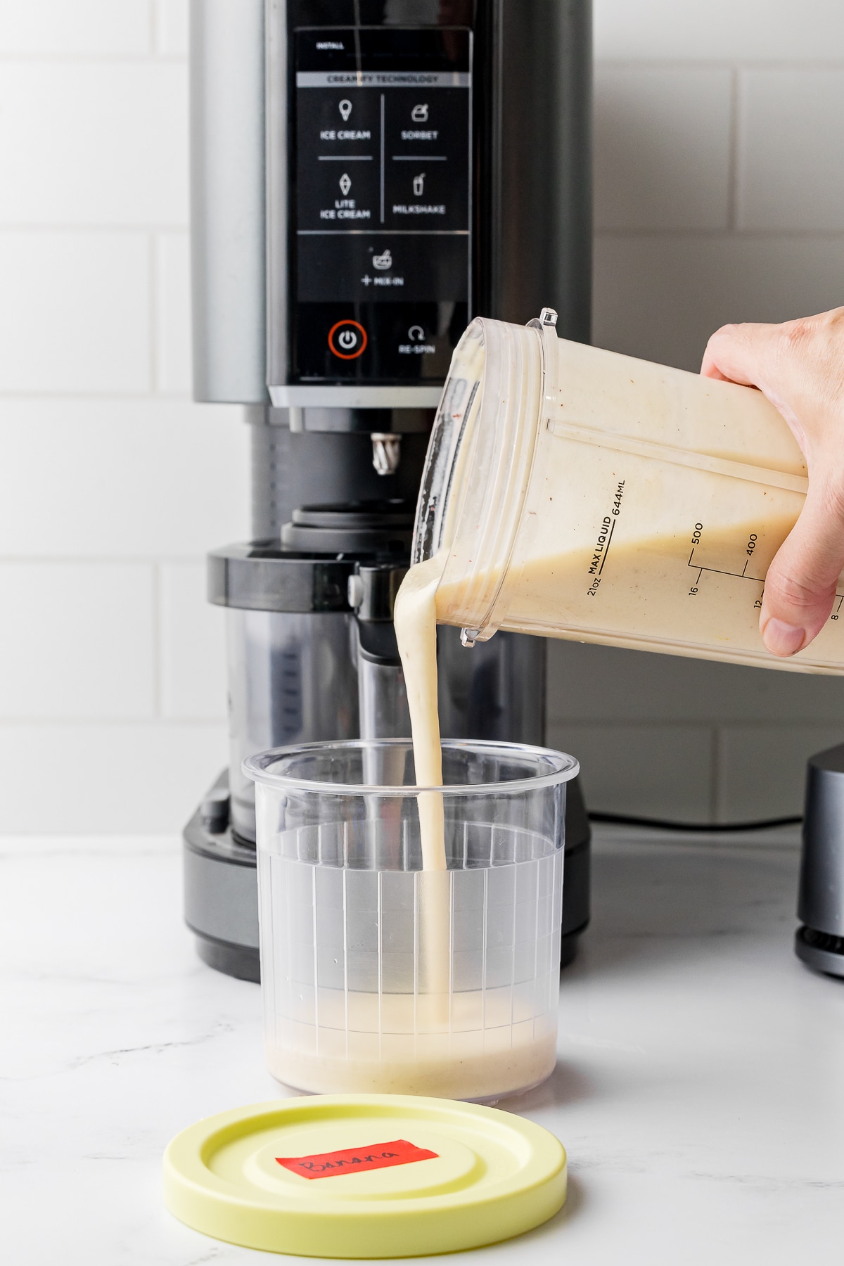 Hand pouring liquid into a Ninja Creami pint with a Ninja machine in the background