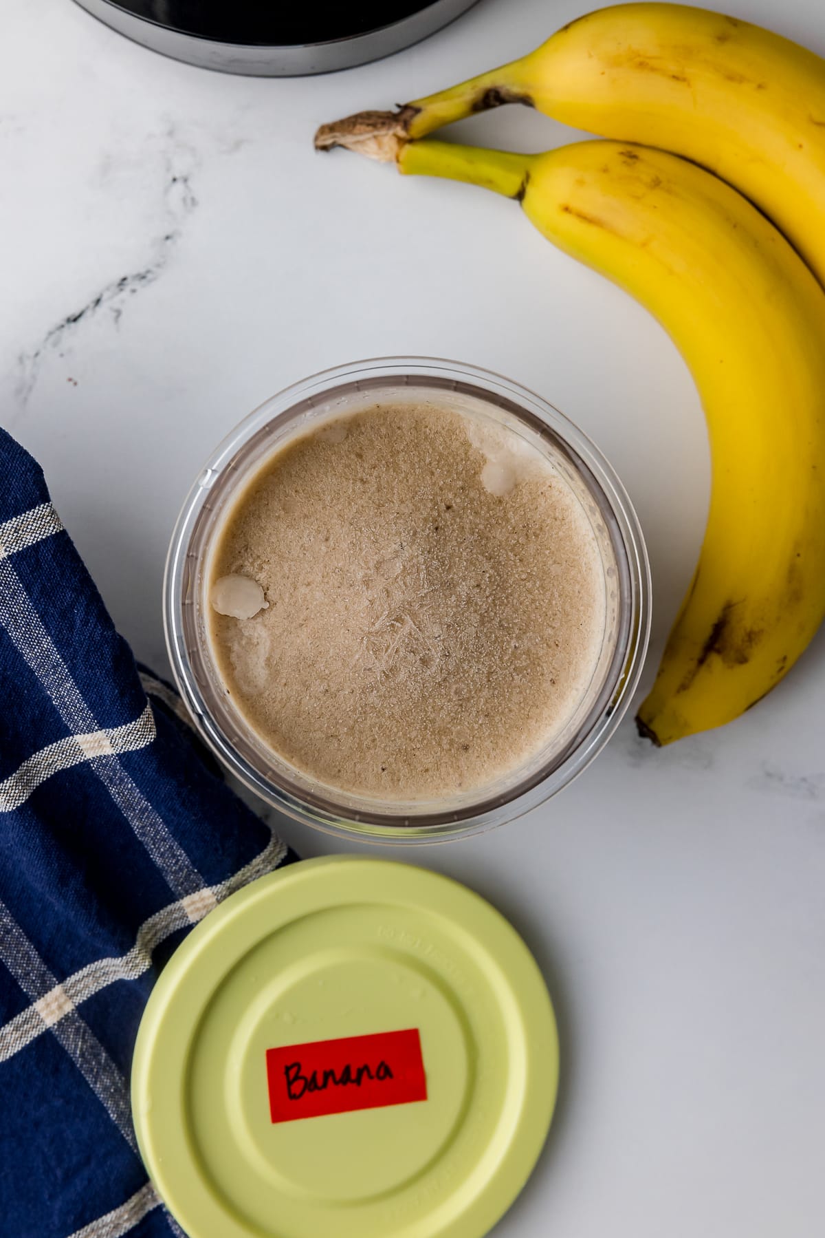 Frozen liquid in a Ninja Creami pint on a white countertop with two bananas and a blue and white kitchen towel