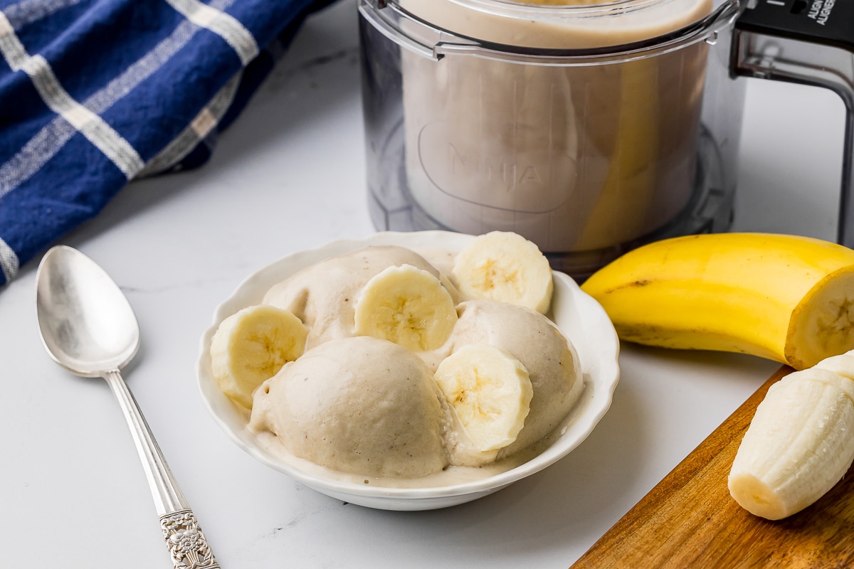 White bowl of ice cream with banana slices on the counter with a sliced banana and a blue and white towel