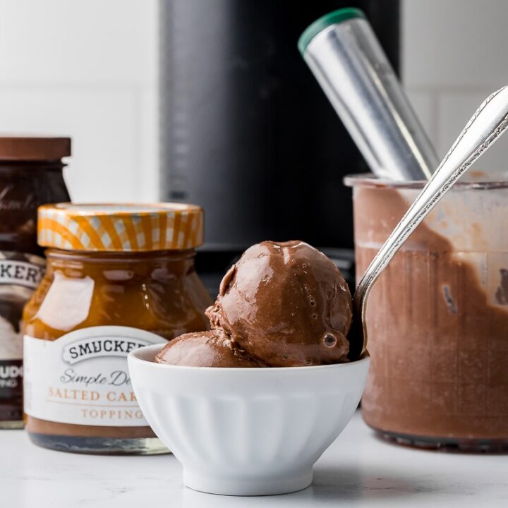 chocolate ice cream in a white bowl with a silver spoon and an ice cream scoop in a ninja creami pint jar