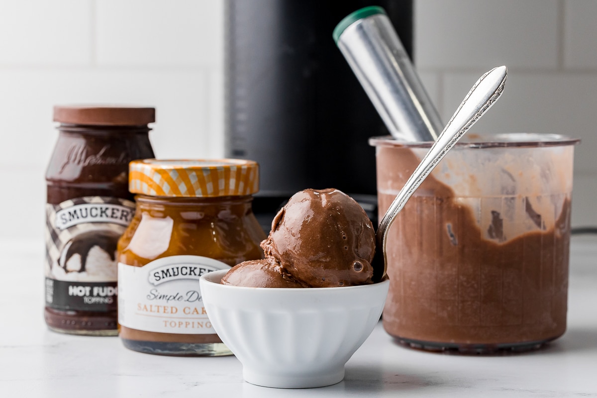 chocolate ice cream in a white bowl with a silver spoon and an ice cream scoop in a ninja creami pint jar