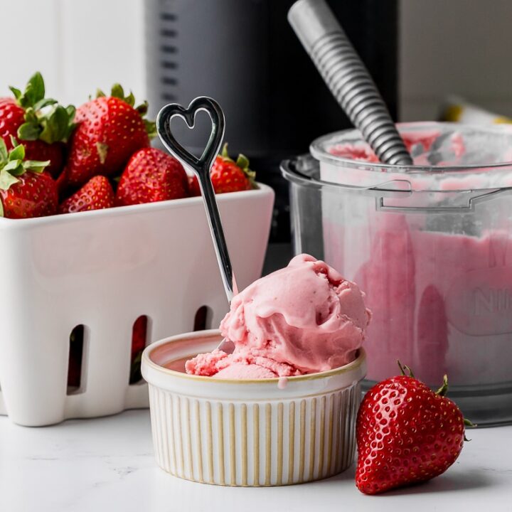 strawberry ice cream in a ramekin with a bowl of strawberries, a ninja pint jar with an ice cream scoop, and a ninja creami in the background