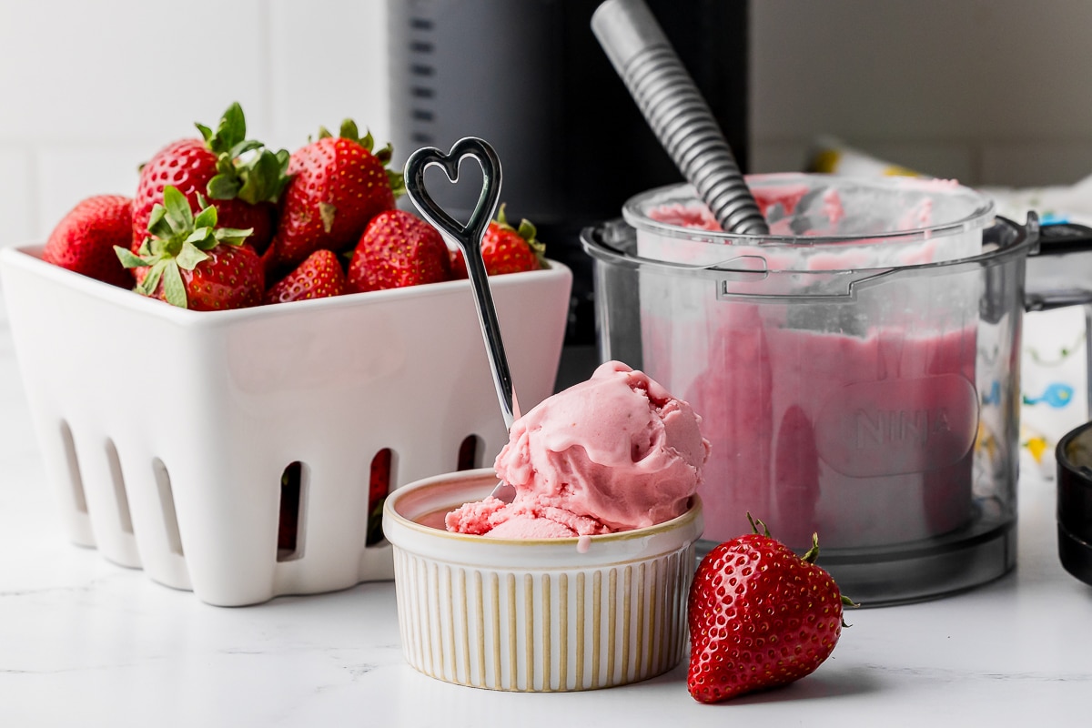 strawberry ice cream in a ramekin with a bowl of strawberries, a ninja pint jar with an ice cream scoop, and a ninja creami in the background