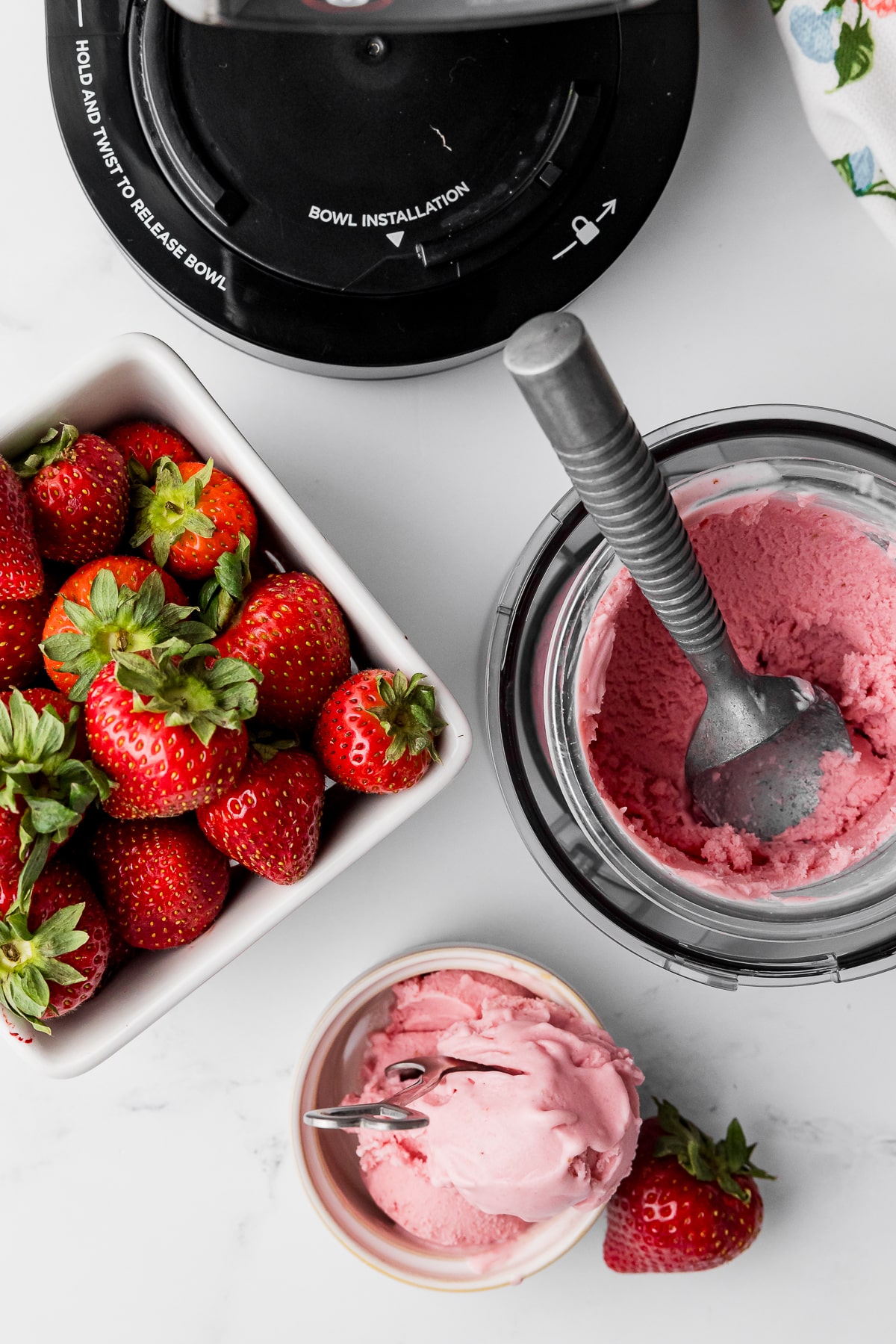 strawberry ice cream in a small bowl with a spoon, an ice cream scoop in a pint jar of ice cream, a basket of fresh strawberries and a ninja creami ice cream machine.