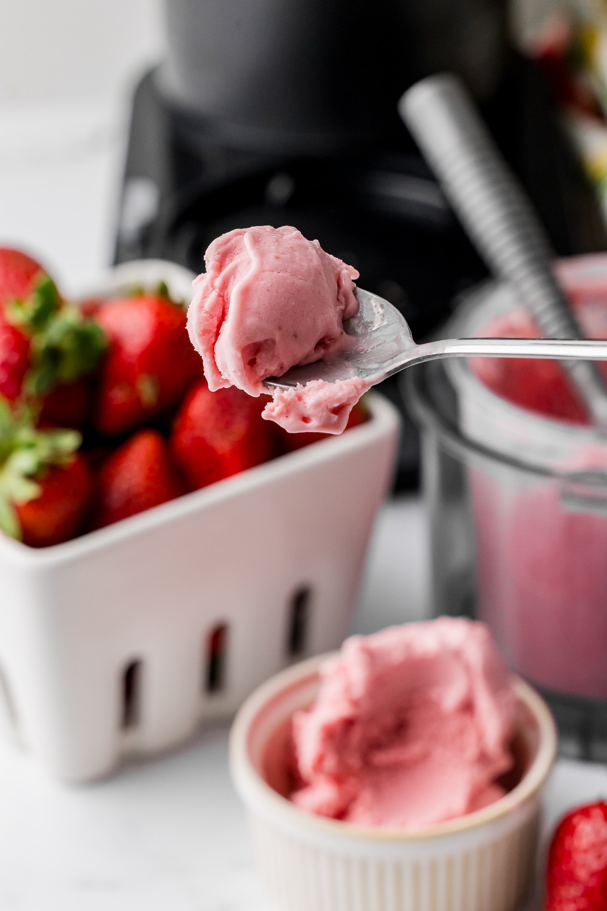 strawberry ice cream on a spoon in front of a basket of fresh strawberries and a ninja creami ice cream machine