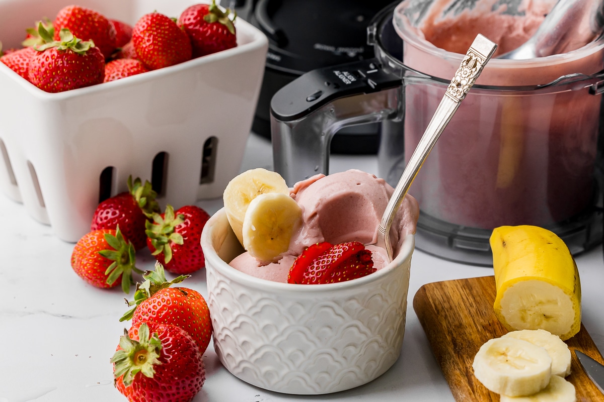 bowl of strawberry banana ice cream with sliced bananas and sliced strawberries with a ninja creami pint jar and a berry bowl full of strawberries