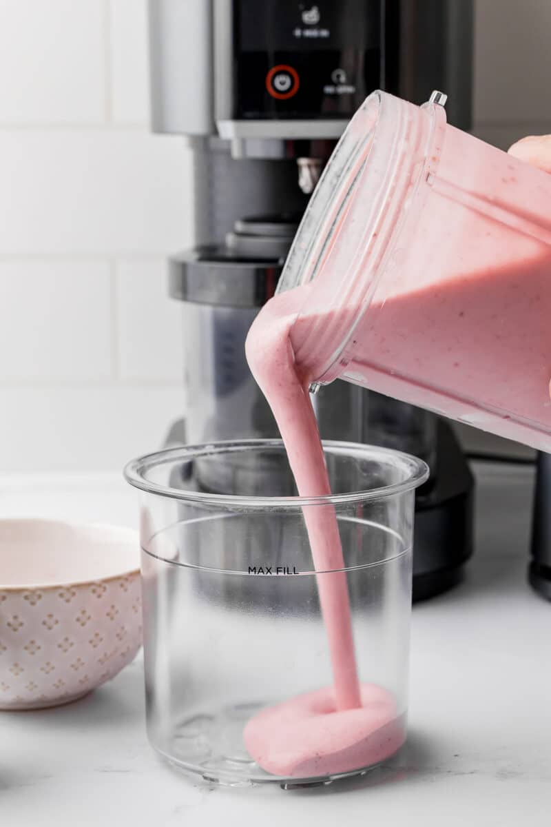 Strawberry banana mixture pouring into a ninja creami pint jar with a ninja creami in the background