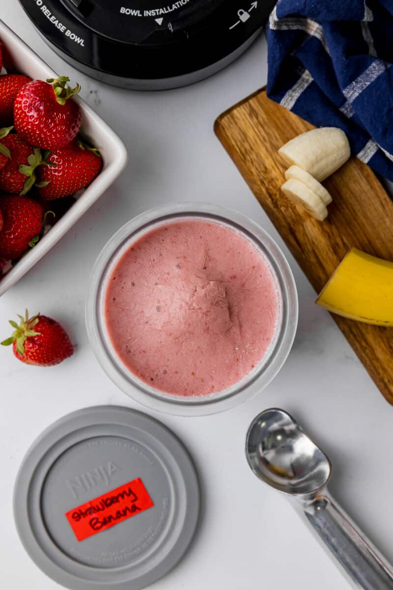 Frozen strawberry banana ice cram in a ninja creami pint jar with fresh strawberries, sliced bananas, and a silver ice cream scooper