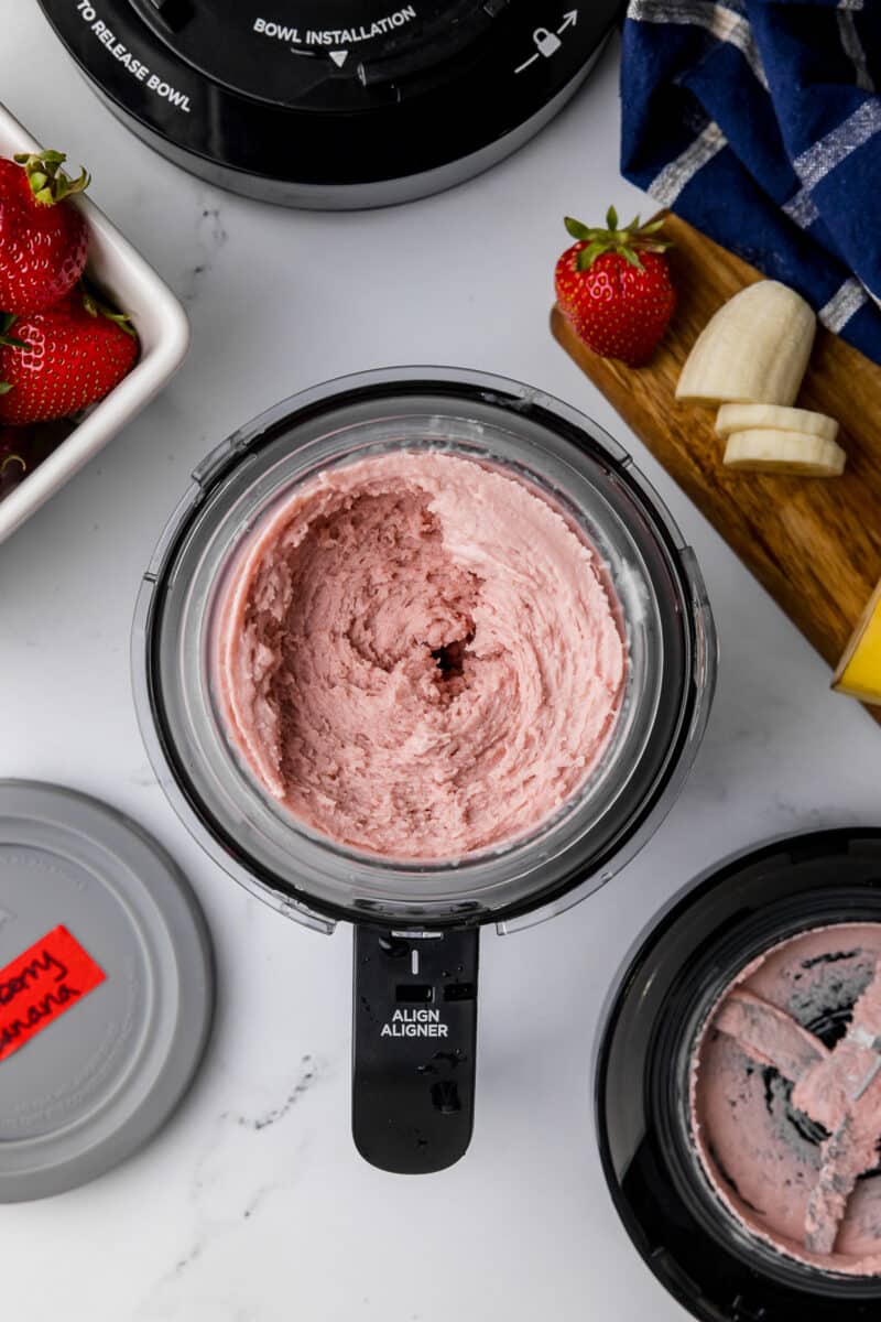 Frozen and spun strawberry banana ice cram in a ninja creami pint jar with fresh strawberries, sliced bananas, and a silver ice cream scooper