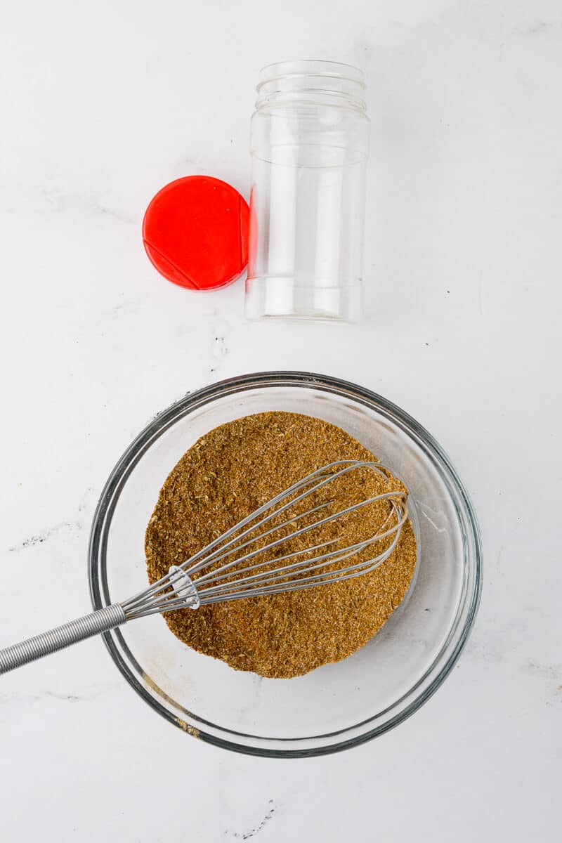 dry mixture of gluten free taco seasoning in a clear bowl with a wire whisk and an empty spice jar and a red lid