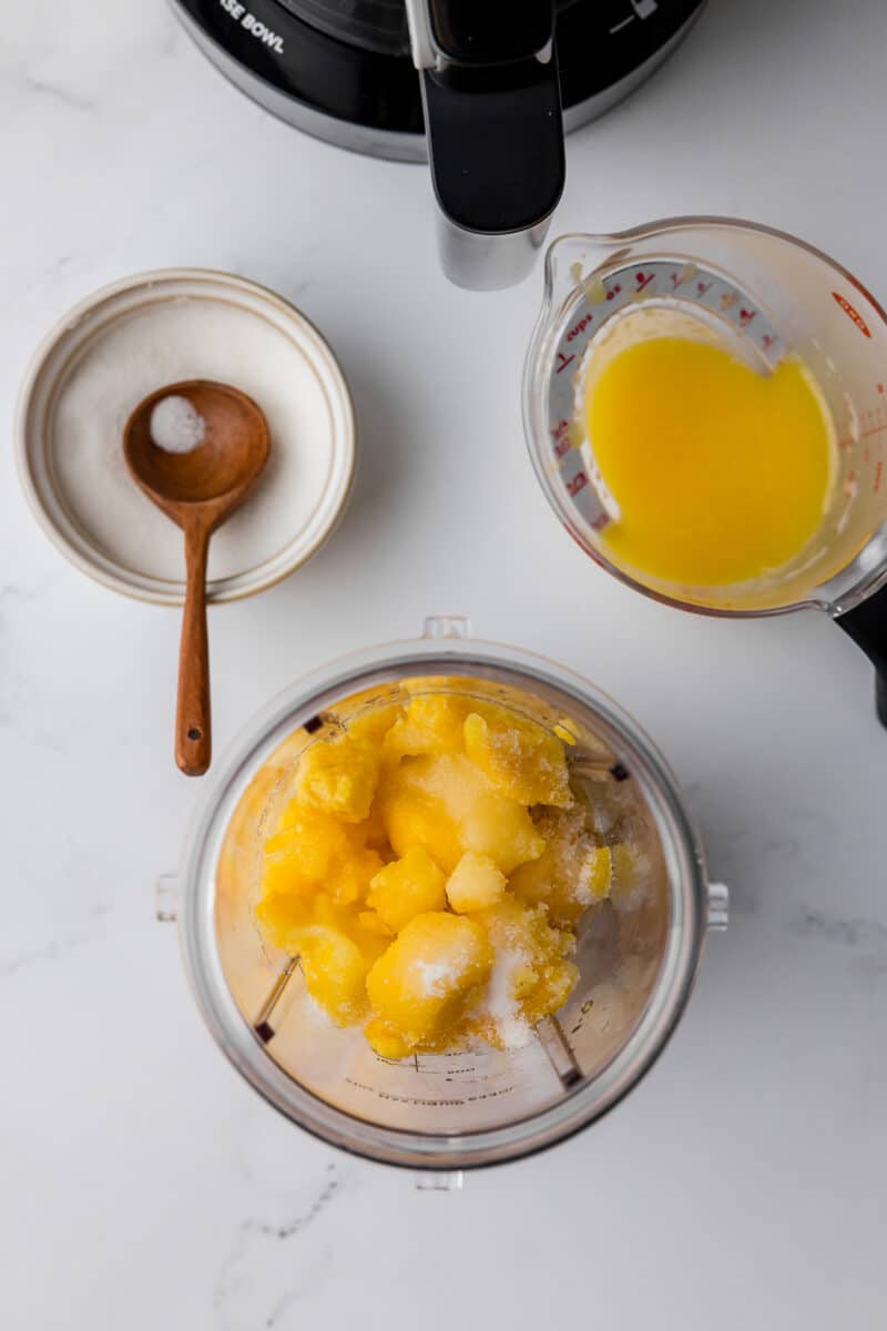 blender with mango chunks inside with sugar with a sugar bowl and a measuring spoon with orange juice