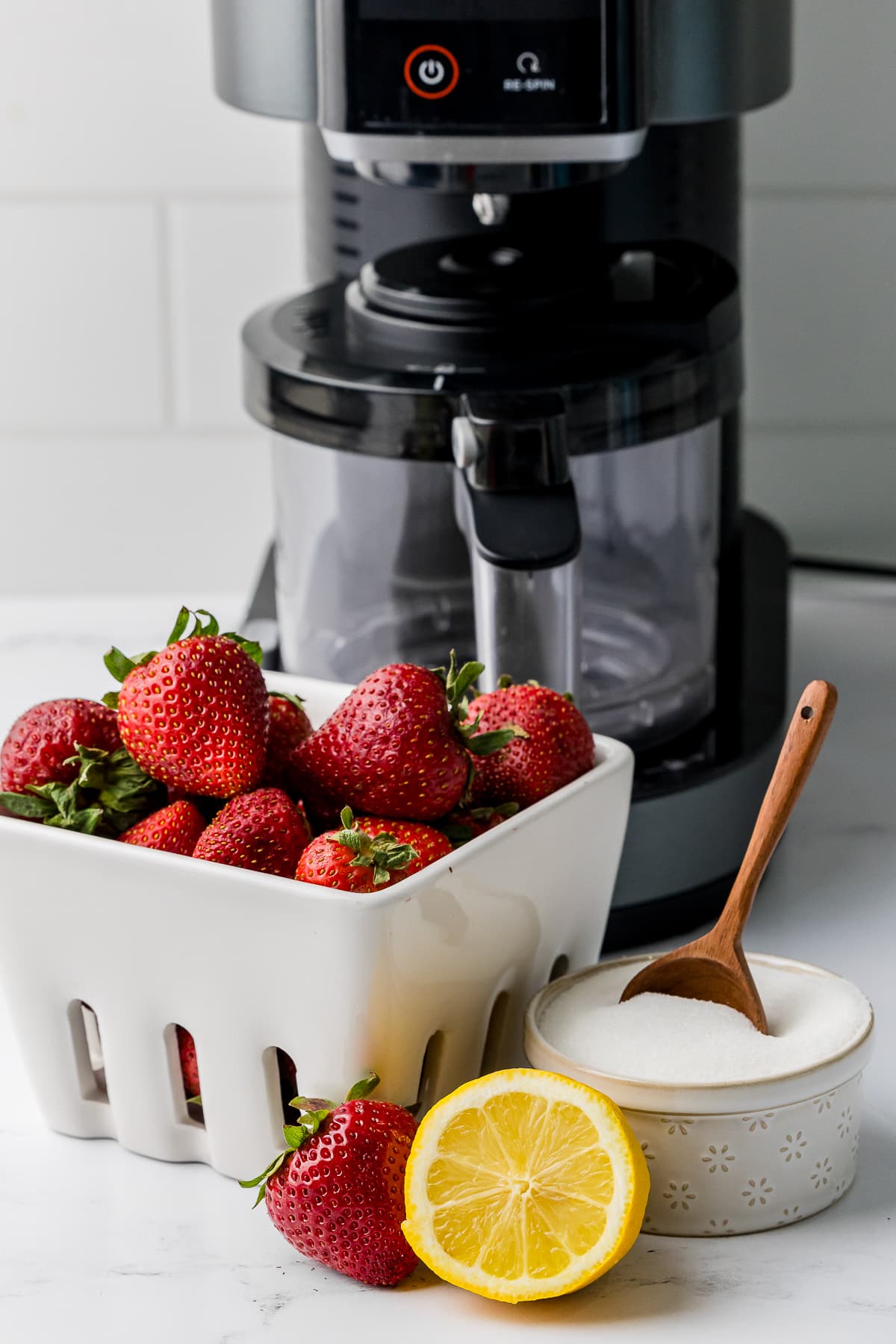 strawberries, half a lemon, sugar in a bowl with a wooden spoon, and a ninja creami ice cream maker