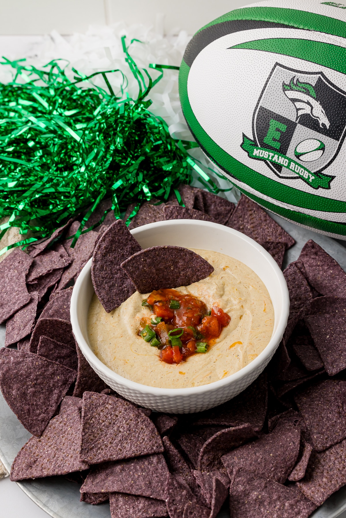 blue corn tortilla chips on a serving plate with green and white pom poms and a rugby ball with cottage cheese queso