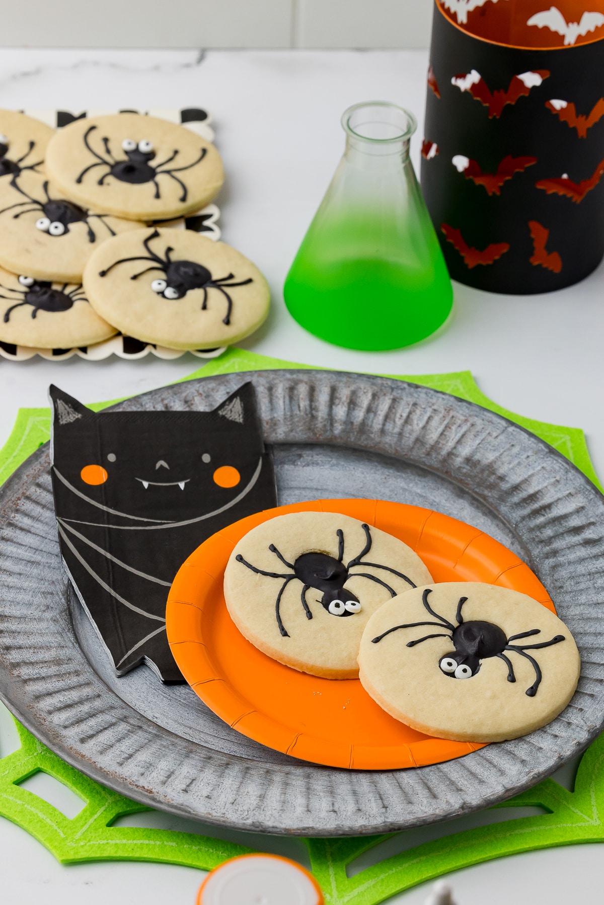 spider sugar cookies on a plate with a bat napkin on a metal plate with a beaker and bat candle holder