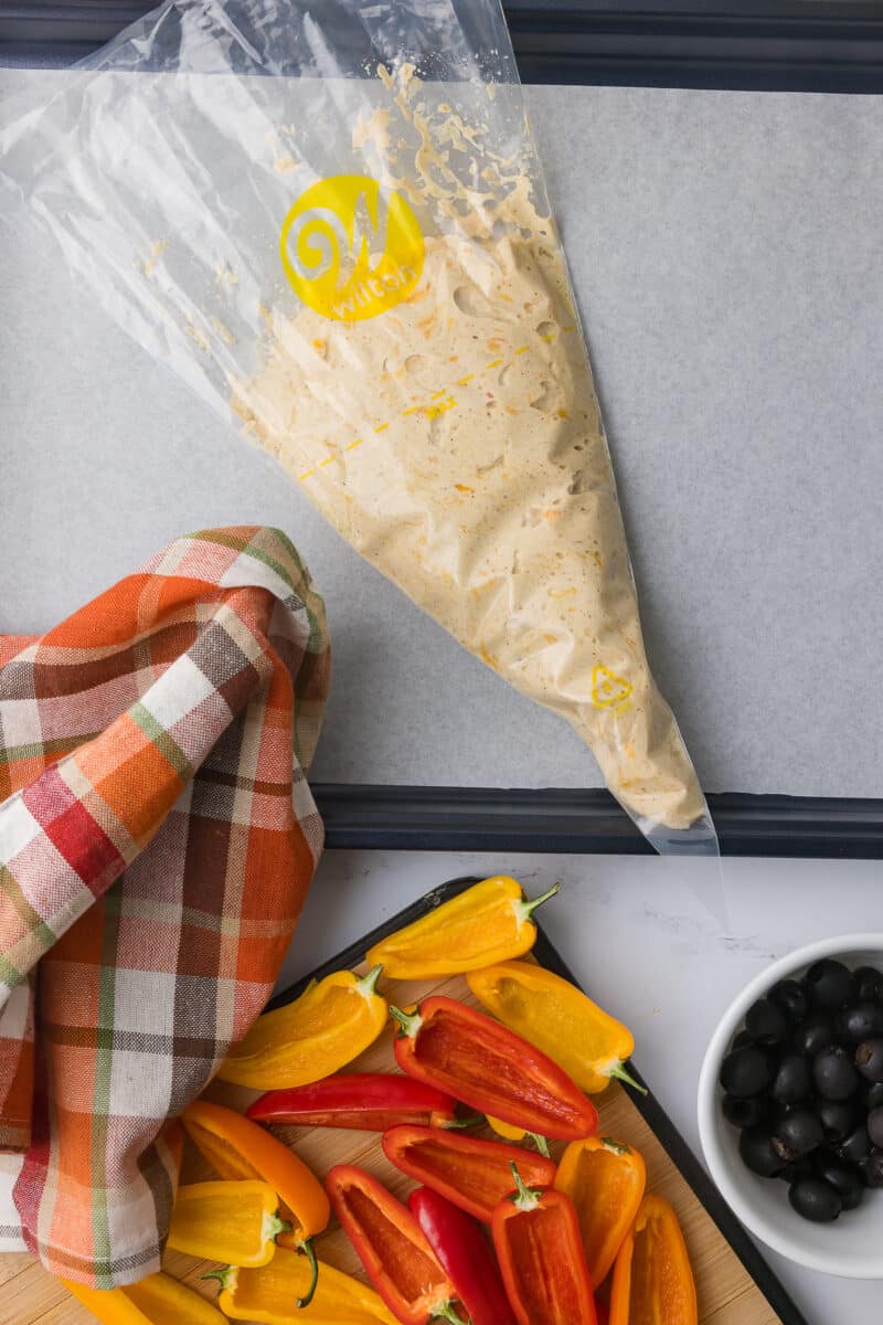 wilton piping bag filled with cottage cheese queso on a wilton baking sheet lined with parchment paper, a festive fall napkin and sliced peppers with olives on a white counter