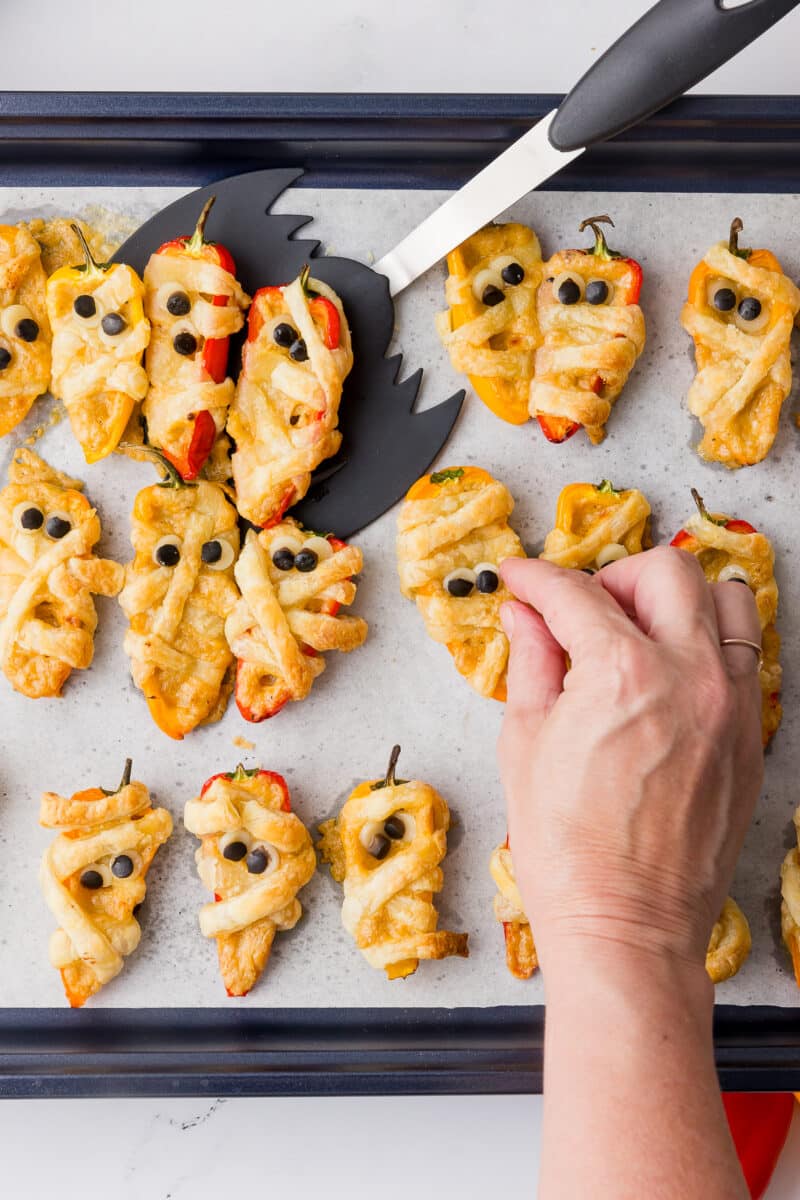 setting olive eyes on baked halloween poppers in a baking sheet lined with parchment paper and a wilton bat spatula