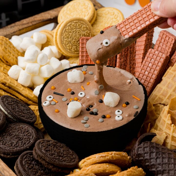 A hand dipping a cookie wafer info a bowl of chocolate hot cocoa dip among a plate of cookies