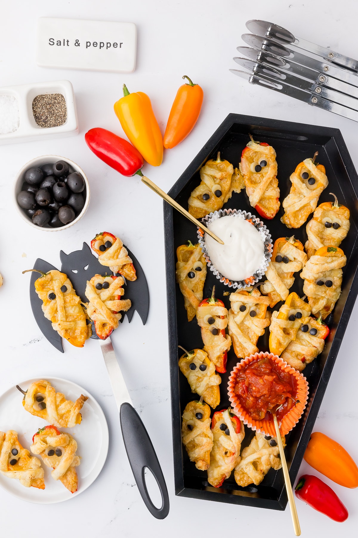 easy halloween poppers in a coffin tray with tri colored peppers, a bowl of olives, and a wilton bat spatula on a white counter