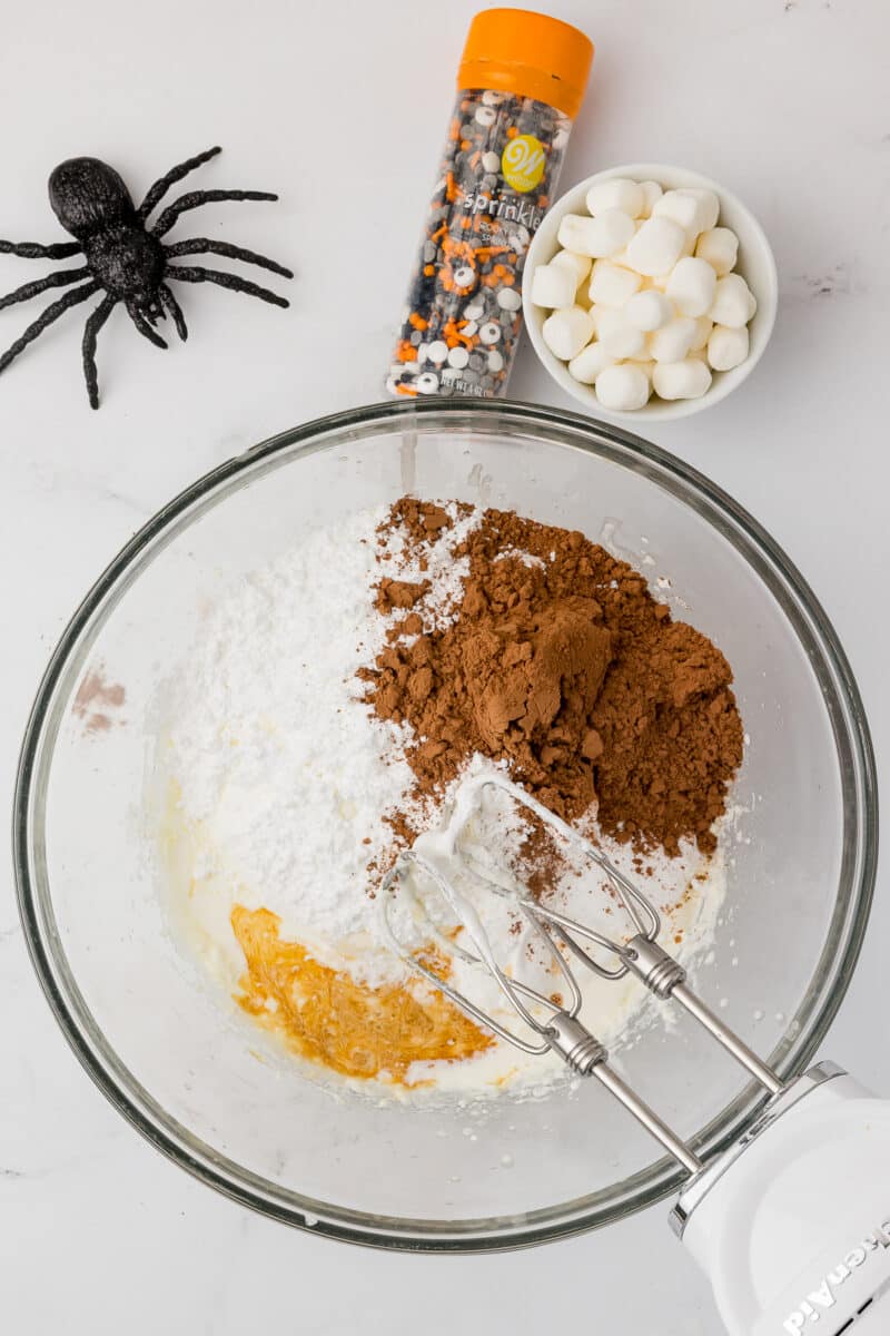 Clear glass bowl full of powdered ingredients with sprinkles and a bowl of marshmallows