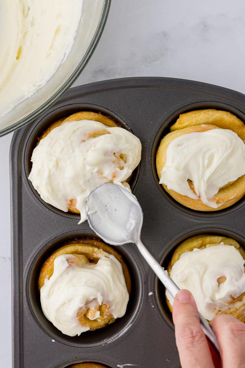 spreading cream cheese frosting on warm lemon rolls still in the muffin pan