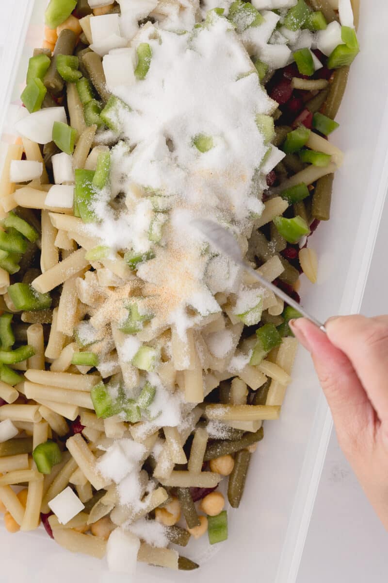 adding sugar and salt to tupperware rectangular dish with beans, onions, and green peppers on a white countertop