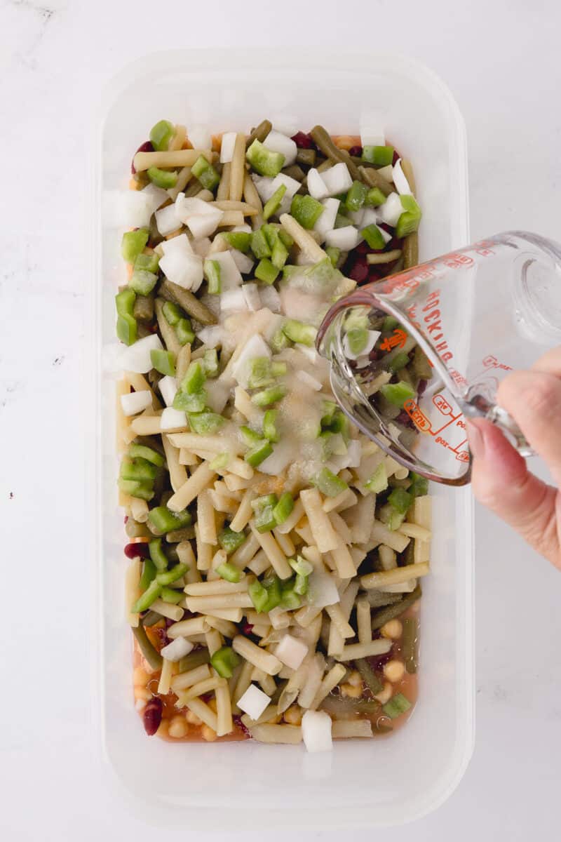adding sugar and salt to tupperware rectangular dish with beans, onions, and green peppers on a white countertop pouring the oil and red wine vinegar