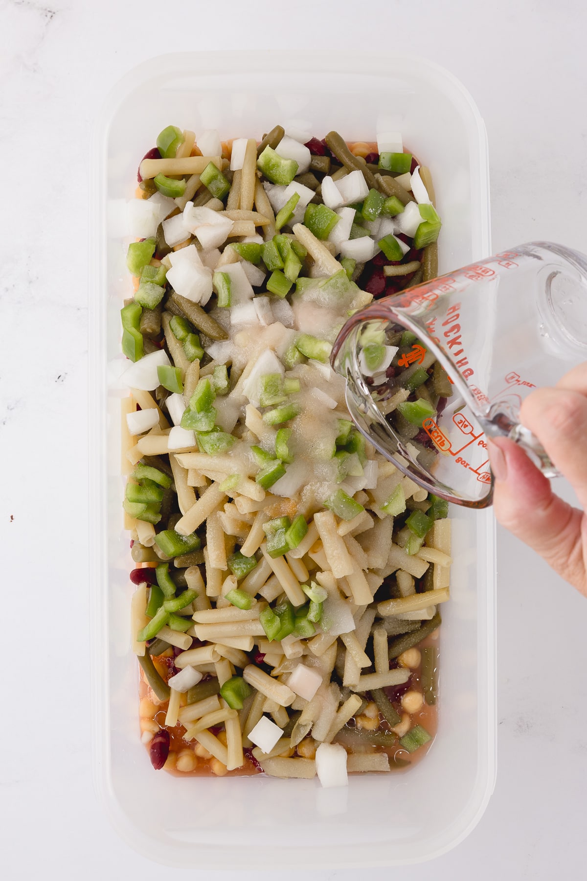 adding sugar and salt to tupperware rectangular dish with beans, onions, and green peppers on a white countertop pouring the oil and red wine vinegar